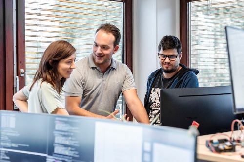 Drei Personen besprechen gemeinsam Inhalte an einem Arbeitsplatz vor mehreren Monitoren.
