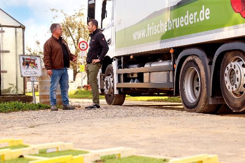 Zwei Personen stehen neben einem Lieferwagen mit der Aufschrift „www.frischebruder.de“ und sprechen miteinander auf einem Hofgelände.