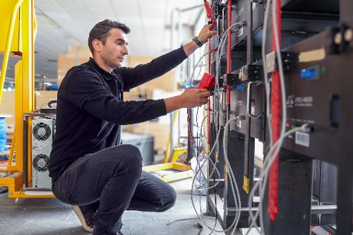 Person kniet vor einem Technikregal und prüft mit einem Messgerät elektrische Verbindungen und Kabel.