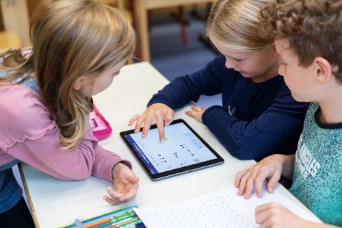 Drei Kinder sitzen gemeinsam an einem Tisch und nutzen ein Tablet mit einer Lernanwendung, während daneben Stifte und ein Arbeitsblatt liegen.