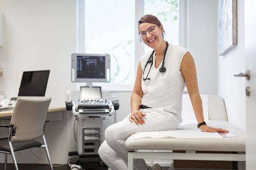 Person in medizinischer Arbeitskleidung sitzt auf einer Untersuchungsliege in einem Behandlungsraum.