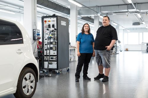 Zwei Personen stehen in einer Halle neben einem Elektrofahrzeug und einem technischen Prüfstand mit angeschlossenen Kabeln und Messgeräten.