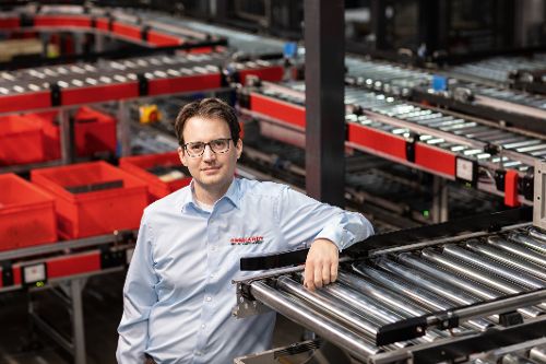 Person steht in einem automatisierten Logistikzentrum neben einer Förderanlage mit Rollenbahnen und roten Transportbehältern.