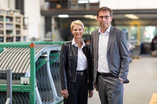 Zwei Personen stehen in einer Produktionshalle neben industriellen Fertigungskomponenten und blicken in die Kamera.