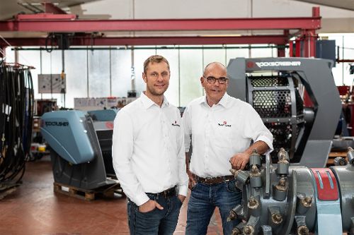 Zwei Personen stehen in einer Produktionshalle vor industriellen Maschinen und technischen Anlagen.