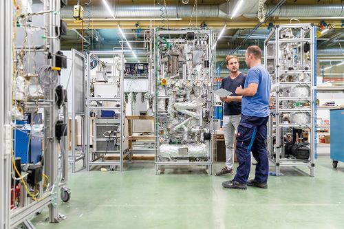 Zwei Personen stehen in einer Produktionshalle vor komplexen technischen Anlagen und besprechen Unterlagen.