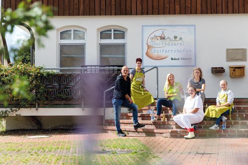 Mehrere Personen sitzen und stehen vor dem Gebäude einer Dorfkäserei und posieren gemeinsam auf einer Treppe.