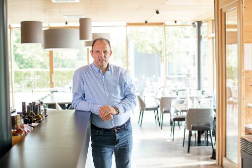 Person steht in einem Gastronomiebereich am Tresen. Im Hintergrund sind Tische, Stühle und große Fenster zu sehen.