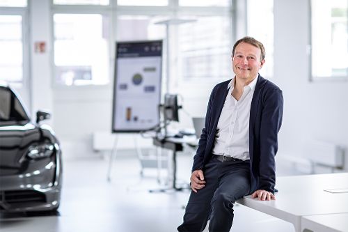 Person sitzt in einer hellen Entwicklungs- oder Forschungsumgebung auf einer Tischkante. Im Hintergrund sind ein Fahrzeug, technische Arbeitsplätze und eine Präsentationstafel zu sehen.