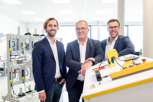Drei Personen stehen in einer technischen Entwicklungs- oder Produktionsumgebung neben einer Anlage mit elektronischen Komponenten und posieren für ein Foto.