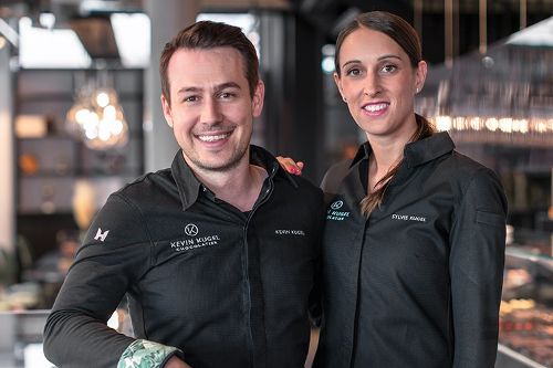 Zwei Personen stehen in einem Gastronomiebetrieb vor einer Verkaufstheke und posieren gemeinsam für ein Foto.