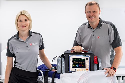 Zwei Personen stehen in einer medizinischen Trainingsumgebung neben einer Patiententrage mit einem medizinischen Überwachungsgerät und posieren für ein Foto.