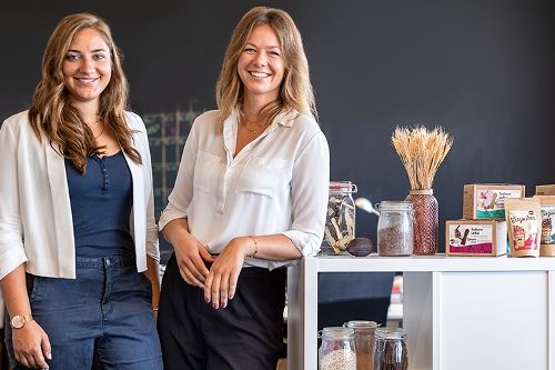 Zwei Frauen stehen in einer Büroumgebung neben einem Regal mit Mustern und Produktverpackungen und posieren für ein Foto.