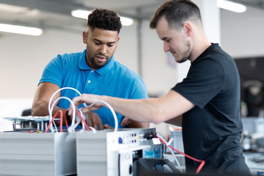 Zwei Personen arbeiten gemeinsam an einer elektrischen Anlage und verbinden Kabel an technischen Komponenten auf einem Arbeitstisch.