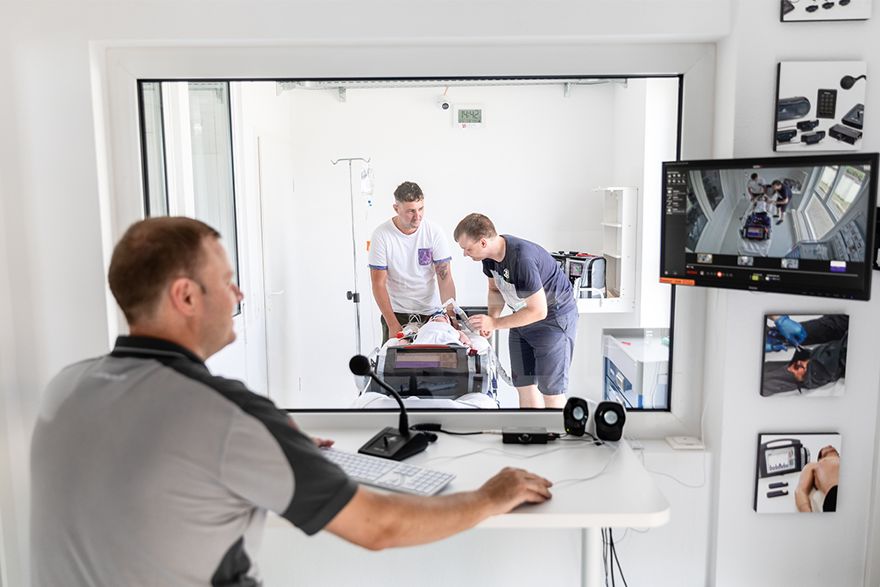 Person beobachtet durch ein Sichtfenster eine medizinische Trainingssituation, unterstützt durch Videoüberwachung.
