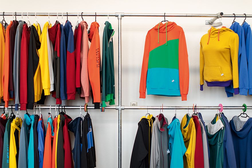 Mehrere farbenfrohe Kapuzenpullover und weitere Kleidungsstücke hängen auf Kleiderstangen in einem Ausstellungs- oder Verkaufsraum.