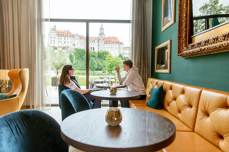 Zwei Personen sitzen in einem stilvoll eingerichteten Café an einem Tisch und unterhalten sich. Durch die großen Fenster ist ein Schlossgebäude im Hintergrund zu sehen.