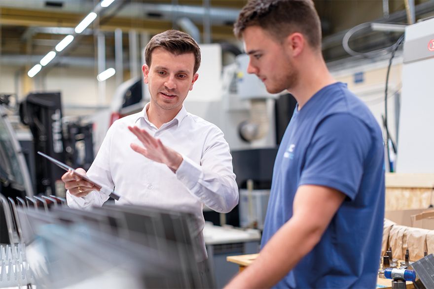 Zwei Personen tauschen sich in einer Produktionsumgebung aus. Im Hintergrund sind technische Anlagen und Arbeitsbereiche zu sehen.
