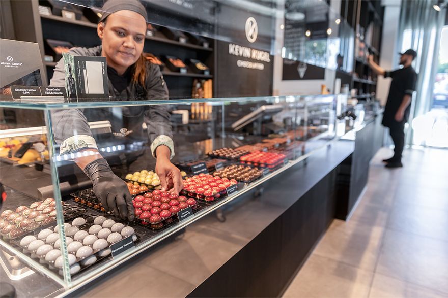 Person arrangiert Pralinen und Schokoladenprodukte in einer Verkaufsvitrine. Im Hintergrund sind weitere Auslagen und Verkaufsflächen eines Fachgeschäfts zu sehen.