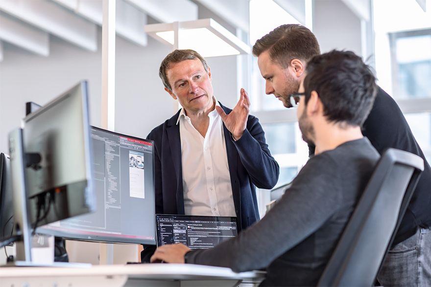 Drei Personen arbeiten gemeinsam an einem Computerarbeitsplatz und besprechen Inhalte auf mehreren Bildschirmen in einer modernen Büroumgebung.