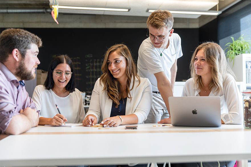 Fünf Personen arbeiten gemeinsam an einem Tisch mit Laptop in einem modernen Büro.