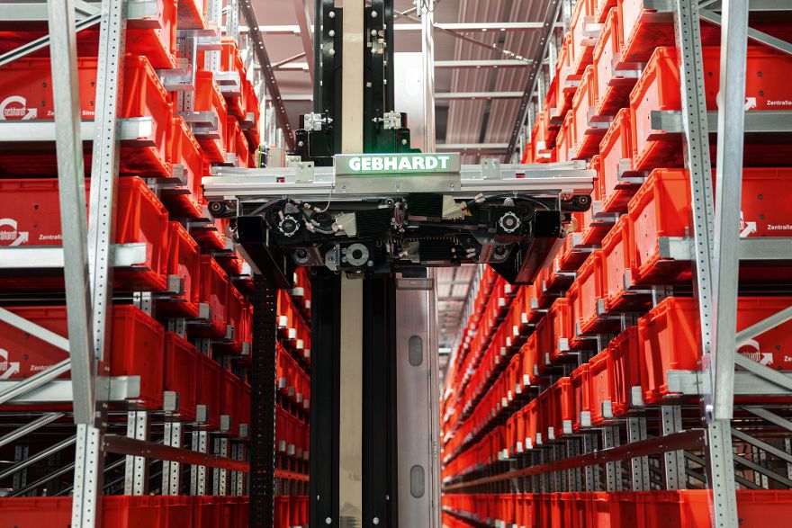 Automatisiertes Kleinteilelager mit langen Regalreihen voller roter Lagerbehälter und einem zentralen Regalbediengerät zur automatischen Ein- und Auslagerung von Waren.