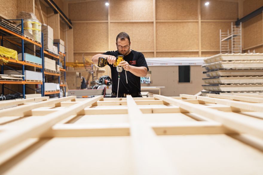 Person verschraubt Holzelemente mit einem Akkuschrauber in einer Produktionshalle, während im Vordergrund ein Holzrahmen auf einer Arbeitsfläche liegt.