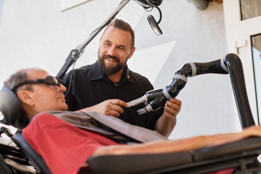Person demonstriert die Bedienung eines technischen Assistenzsystems mit einem robotischen Arm, während eine weitere Person das System nutzt.
