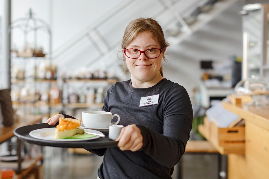 Person steht in einem Café und hält ein Tablett mit einer Tasse Kaffee und einem kleinen Dessert nach vorne.