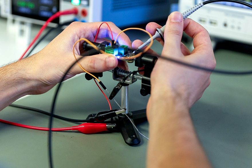 Hände verbinden elektronische Bauteile mit Kabeln auf einer kleinen Halterung in einem Laboraufbau.