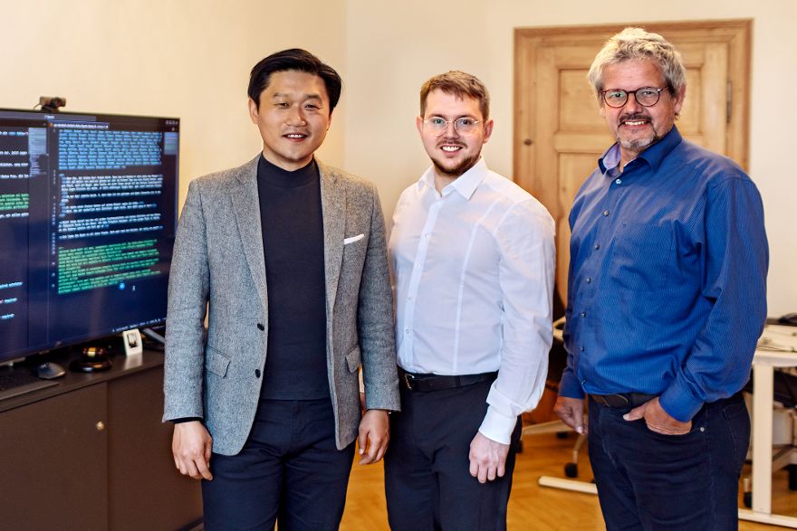 Drei Männer stehen in einem Büro vor einem großen Bildschirm mit Codeanzeige.