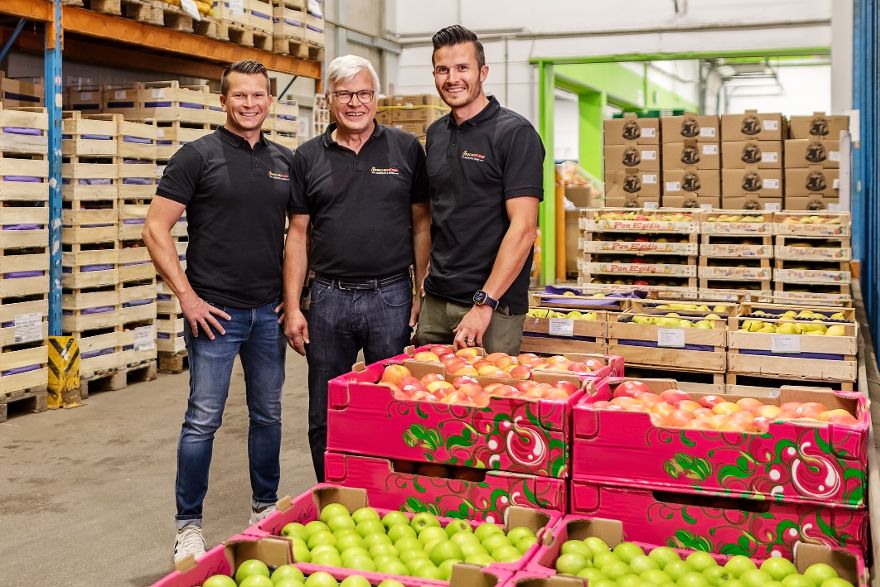 Drei Männer stehen in einem Lager zwischen gestapelten Kisten und Paletten gefüllt mit Obst.