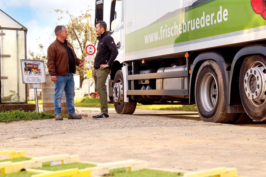 Zwei Personen stehen neben einem Lieferwagen mit der Aufschrift „www.frischebruder.de“ und sprechen miteinander auf einem Hofgelände.
