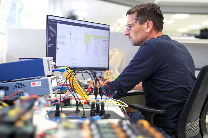 Person arbeitet an einem Arbeitsplatz mit elektronischen Komponenten und schaut auf einen Monitor mit Schaltplänen.