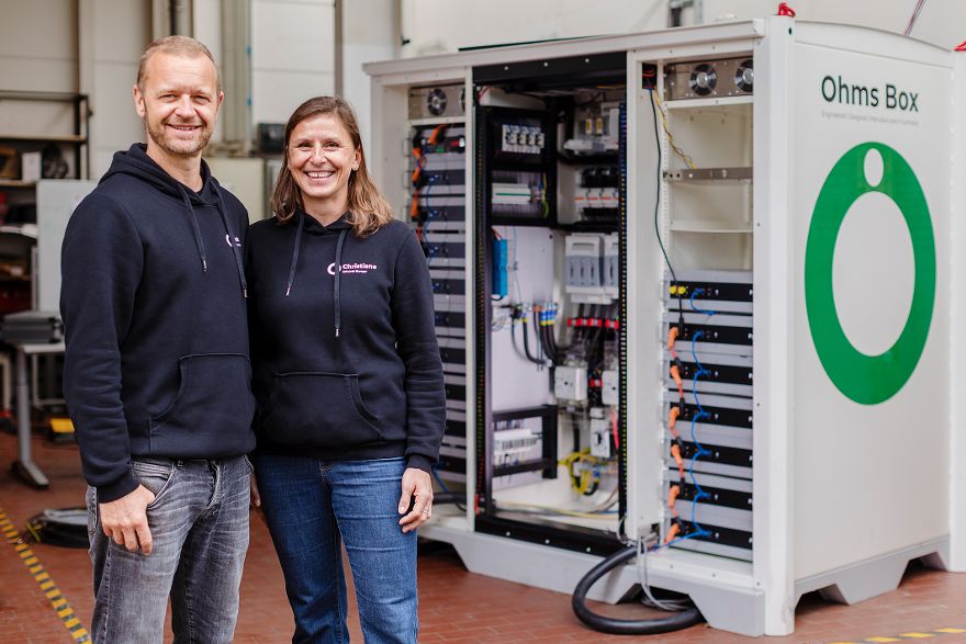 Zwei Personen stehen in einer Werkhalle neben einem geöffneten Technikschrank mit sichtbarer Elektronik.