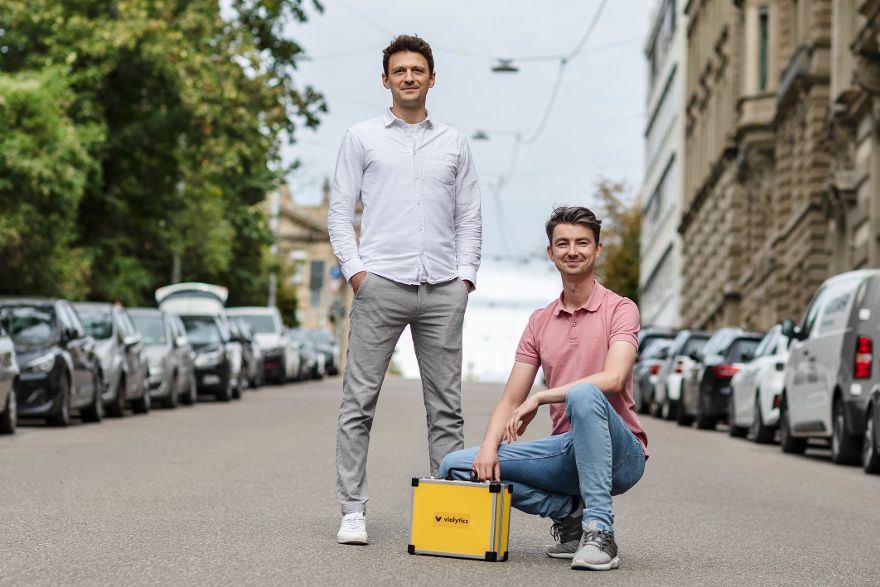 Zwei Personen stehen auf einer Straße zwischen geparkten Autos, während eine Person einen gelben Koffer in der Hand hält und die andere daneben steht.