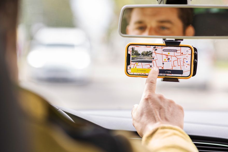 Hand bedient ein Smartphone mit Navigationskarte, das im Auto an der Windschutzscheibe befestigt ist, während im Rückspiegel ein Gesicht sichtbar ist.