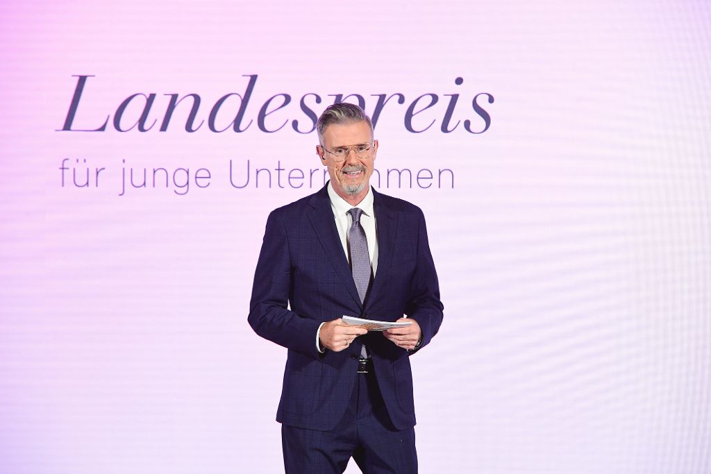 Mann mit Moderationskarten steht in Anzug auf Landespreis-Bühne.