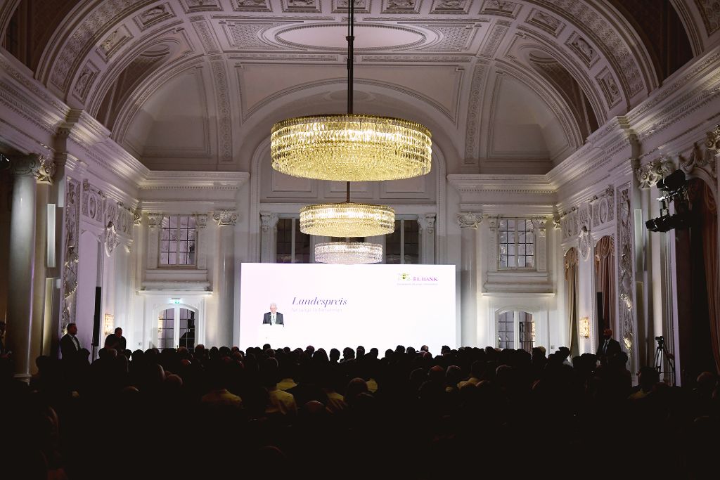 Publikum sitzt in einem festlichen Saal mit großen Kronleuchtern und blickt auf eine Bühne mit einer Präsentation zum „Landespreis für junge Unternehmen“.