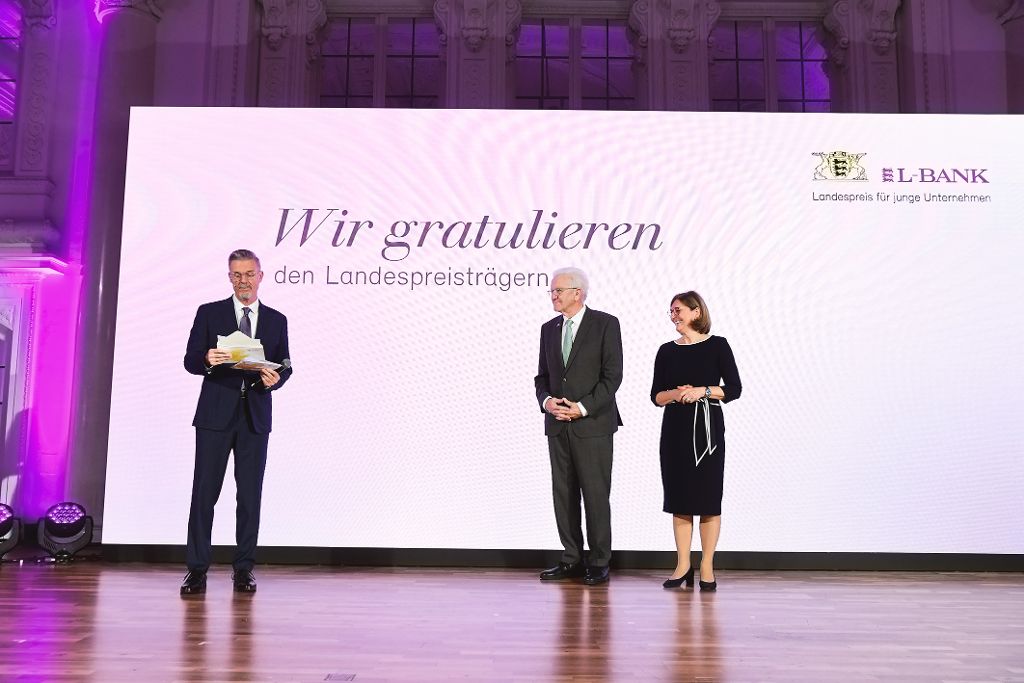 Drei Personen stehen auf einer Bühne vor einer großen Leinwand mit der Aufschrift „Wir gratulieren den Landespreisträgern“ im Rahmen der Verleihung des Landespreises für junge Unternehmen.