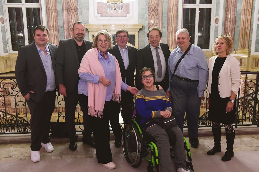 Gruppenfoto mehrerer Personen in einem festlich ausgestatteten Gebäude. Eine Person sitzt im Rollstuhl, die übrigen Personen stehen daneben vor einem Geländer im Innenbereich.