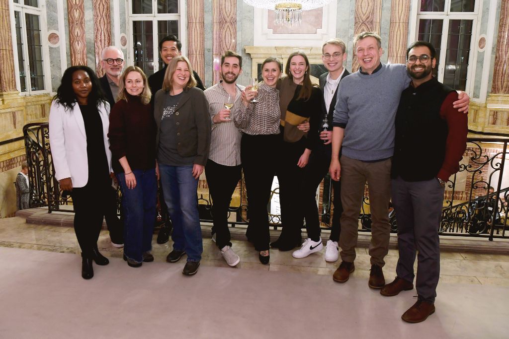 Gruppenfoto mehrerer Personen in einem festlich ausgestatteten Innenraum. Die Gruppe steht vor einem Geländer und posiert gemeinsam für die Kamera, einige halten Gläser in der Hand.