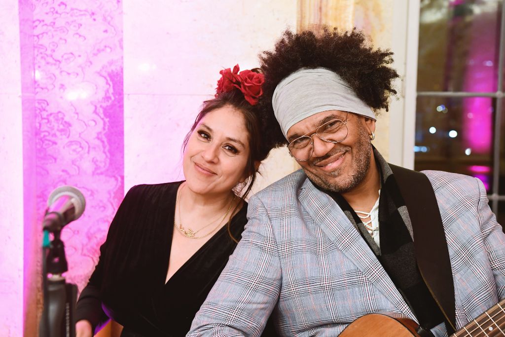 Zwei Musiker sitzen nebeneinander bei einer Veranstaltung. Eine Person hält eine Gitarre, im Vordergrund ist ein Mikrofon zu sehen, im Hintergrund eine dekorativ beleuchtete Innenraumkulisse.