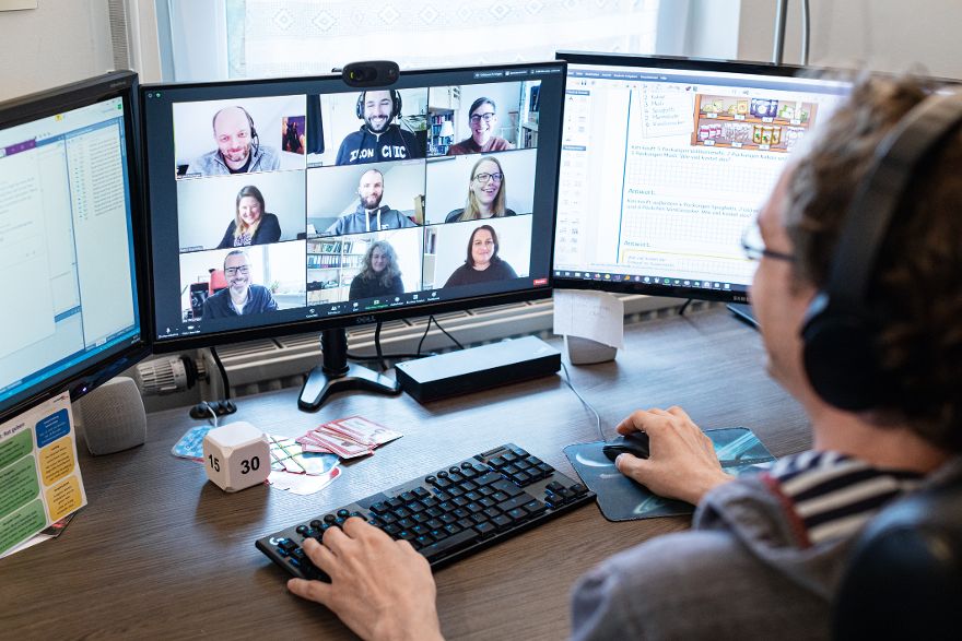 Person sitzt an einem Schreibtisch mit drei Monitoren und nimmt an einer Videokonferenz mit mehreren Teilnehmenden teil.